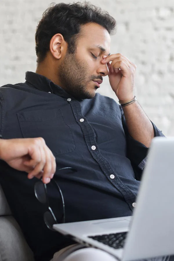 Adrenal-Fatigue-Treatment A middle-aged man with a laptop closing his eyes and rubbing his nose. He needs treatment for Adrenal Fatigue Syndrome from AZENA Health & Longevity in Gainesville.