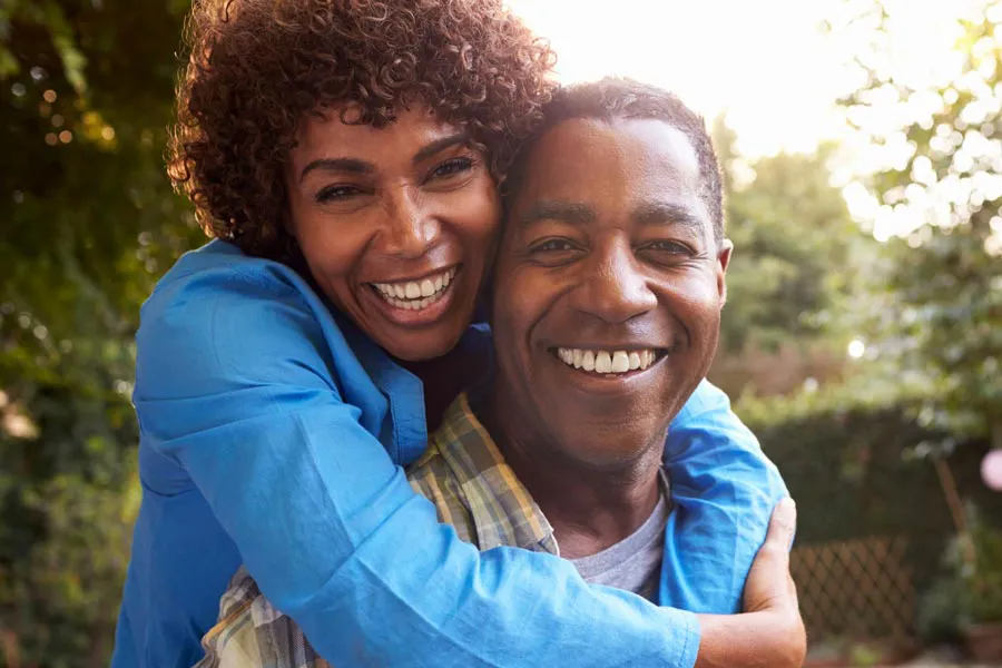 Autoimmune-Doctor A middle-aged African American couple outside, the woman embracing the man from behind pressing her cheek to his, both are smiling due to successful Autoimmune Disorder treatment from AZENA Health & Longevity in Gainesville.