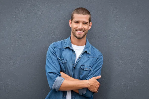 Hormones-for-Men-Doctor A man in a denim shirt stands smiling against a gray-blue wall, pleased with his testosterone hormone therapy from AZENA Health & Longevity in Gainesville.