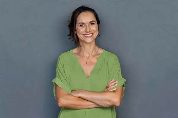 Hormones-for-Women-Doctor A woman in a green shirt stands smiling against a gray-blue wall, pleased with her hormone therapy from AZENA Health & Longevity in Gainesville.
