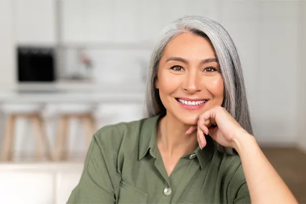 Menopause-Clinic An asian woman with long gray hair and a green shirt sitting and smiling on her couch, happy with her menopause care from [NAME} in Gainesville.