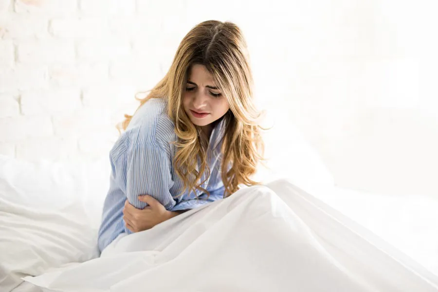PMS-Therapy A woman with long blonde hair sits in her bed holding her stomach in pain, considering getting care for PMS from AZENA Health & Longevity in Gainesville.