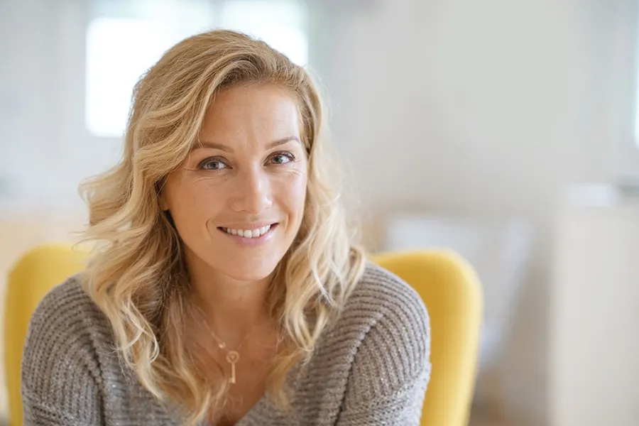 Perimenopause-Doctor A blonde woman wearing a a gray sweater sits inside in a yellow chair, smiling and happy with her perimenopause treatment from AZENA Health & Longevity in Gainesville.